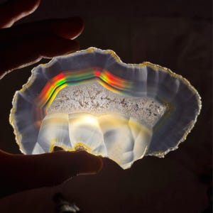 May include: A translucent, polished agate geode slice, showcasing a spectrum of colors including red, yellow, and green. The natural stone features a crystalline center and a layered, banded pattern, with a hand holding the geode.