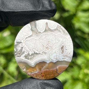 May include: A circular polished gemstone cabochon featuring intricate white and translucent gray banding patterns with a distinct brown earthy base layer. The stone displays organic, cloud-like formations and concentric circular details throughout.