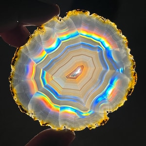 May include: A translucent, circular agate slice with concentric bands of color. The agate features a rainbow iridescence effect, with bands of orange, yellow, green, and blue. The outer edge is a dark brown color. The agate is held between two fingers.