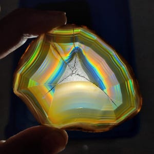 May include: A translucent, polished agate slice with a rainbow of colors. The stone features concentric bands of yellow, green, and blue, with a central, lighter area. Fine cracks are visible across the surface, adding to its natural beauty.