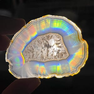 May include: A translucent, irregularly shaped agate slice backlit to reveal concentric bands of white and gray stone. The light creates a vibrant, prismatic rainbow effect along the inner edges, surrounding a textured, crystalline center.