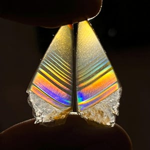 Puede incluir: Dos gemas triangulares iridiscentes con un arcoíris de colores, incluyendo amarillo, naranja, azul y morado. Las gemas tienen un borde rugoso y se presentan sobre un fondo oscuro.