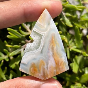 Könnte beinhalten: Ein dreieckiger Edelstein mit einem einzigartigen Muster aus weißen, gelben und hellblauen Wirbeln. Der polierte Stein hat ein natürliches, organisches Aussehen mit einer kleinen Höhle auf einer Seite. Der Edelstein wird in einer Hand gehalten.