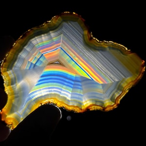 May include: A translucent agate slice featuring concentric bands of white, blue, and orange. The mineral specimen displays a vibrant rainbow-like spectrum of light passing through its crystalline structure, with a jagged, irregular outer edge.
