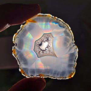 May include: A translucent, polished agate slice with a central, geode-like cavity. The stone displays a spectrum of rainbow colors refracting light. The outer edge has a golden-brown hue, and the overall shape is irregular.