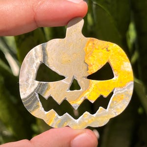 May include: A carved pumpkin-shaped pendant with a spooky face. The stone has a mix of colors, including yellow, gray, and white. The pendant is held up by a person's fingers against a green background.