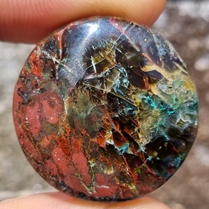 May include: A round, polished stone with a unique, swirling pattern of red, brown, green, and blue colors. The stone has a smooth, glossy finish.