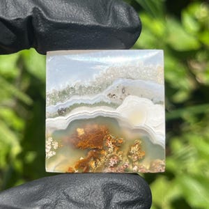 May include: A square-cut polished agate gemstone featuring translucent white and grey banding, with internal moss-like inclusions in shades of rust-orange, brown, and green, displaying a smooth, flat surface with visible crystalline textures.