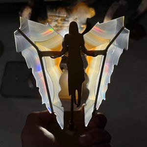 May include: A decorative ornament featuring a dark silhouette of a figure with outstretched arms positioned in front of two translucent, fan-shaped agate slices. The stone slices display banded patterns in white, cream, and orange with iridescent light.