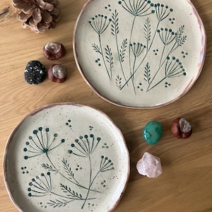 Może przedstawiać: Dwa ręcznie robione ceramiczne talerze z kremową podstawą i ręcznie malowanym botanicznym wzorem w odcieniach zieleni. Talerze są okrągłe z nakrapianą krawędzią. Elementy dekoracyjne, takie jak szyszka, kamienie i kasztany, są ułożone wokół talerzy.