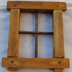 Könnte beinhalten: Ein quadratischer Holzrahmen mit einem Metallgitter im rustikalen Stil. Der Rahmen besteht aus hellbraunem, verwittertem Holz mit sichtbarer Maserung und Nagelköpfen. Das Metallgitter besteht aus vier sich kreuzenden, rostbraunen Stäben, die ein fensterartiges Design bilden.