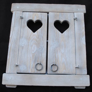 Könnte beinhalten: Ein weiß gestrichener Holzfensterladen mit zwei herzförmigen Ausschnitten. Der Laden ist geschlossen und hat zwei Metallringe an jeder Seite.