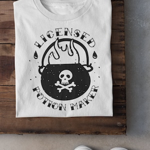 May include: White t-shirt with a black and white graphic of a cauldron with flames and a skull and crossbones. The text "Licensed Potion Maker" is written around the cauldron.