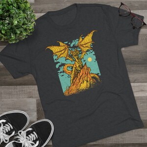 Könnte beinhalten: Ein dunkel-helles graues T-Shirt mit einem Aufdruck eines gelben Drachen mit orangefarbenen Flammen auf einem blauen und grünen Hintergrund.