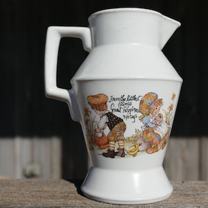 May include: White ceramic jug with a handle and spout. The jug features a colourful illustration of children and the text "From the littlest things great happiness springs". The jug is on a wooden surface.