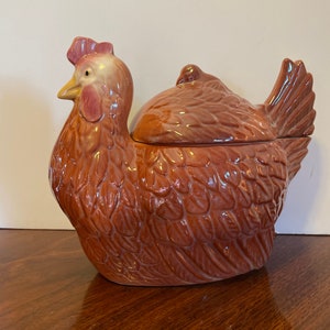 May include: A ceramic hen-shaped container with a lid. The container is primarily brown with a reddish-brown comb and wattle. The design features textured details to resemble feathers. The lid is shaped to fit the hen's back.