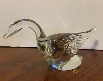 May include: A clear glass figurine of a goose with a curved neck and detailed wing design. The sculpture is transparent, allowing light to pass through, and sits on a wooden surface. The goose is a decorative item.
