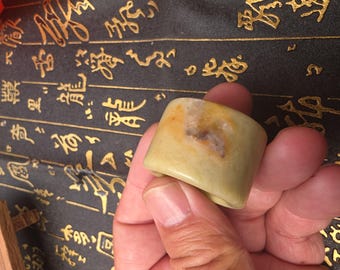 May include: A light green jade ring with a brown and yellow mottled pattern. The ring is being held by a hand.