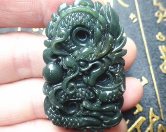 Op de afbeelding: Een donkergroene jade hanger gegraveerd in de vorm van een draak. De draak is ingewikkeld gedetailleerd met schubben en klauwen. De hanger heeft een gat aan de bovenkant voor een koord of ketting.