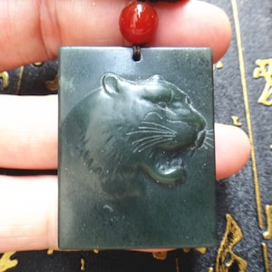 Puede incluir: Un colgante de jade verde oscuro con un diseño de cabeza de tigre tallado. El colgante tiene forma cuadrada y tiene una cuenta de ágata roja en la parte superior.