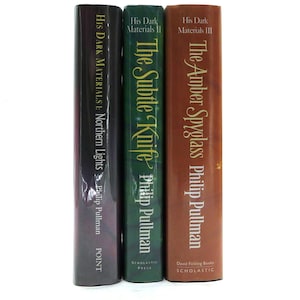 Peut inclure: Trois livres reliés de la série "À la croisée des mondes" de Philip Pullman. Les couvertures sont de couleurs marron, verte et violet foncé. Les titres incluent "Les Royaumes du Nord", "La Tour des Anges" et "Le Miroir d'ambre."