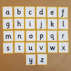 May include: Alphabet flashcards featuring lowercase letters a through z. Each white card has a yellow border and a single black letter. The cards are arranged on a brown surface, ideal for educational use.