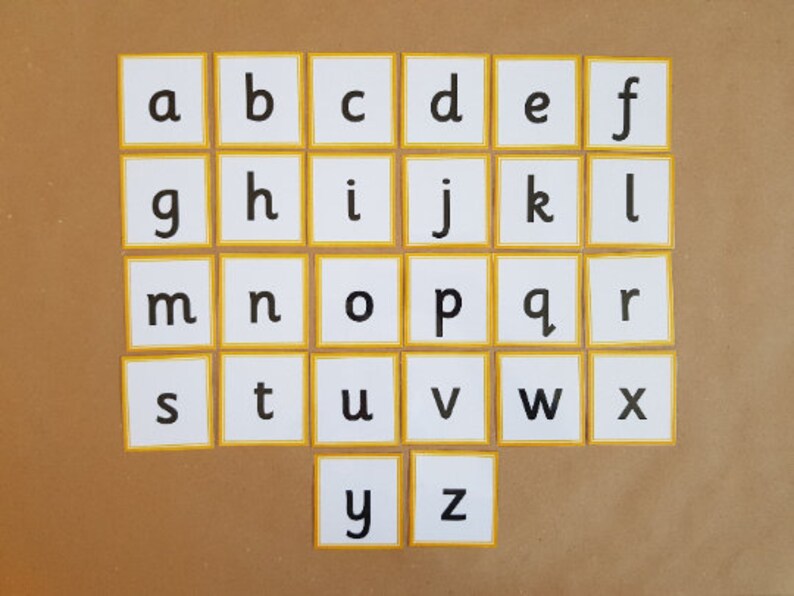May include: Alphabet flashcards featuring lowercase letters a through z. Each white card has a yellow border and a single black letter. The cards are arranged on a brown surface, ideal for educational use.