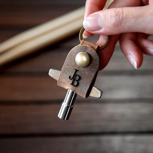 Op de afbeelding: Een bruine leren drum key houder met de initialen "JB" gegraveerd op de voorkant. De houder heeft een metalen drum key bevestigd en een messing klinknagel. Twee houten drumstokken staan op de achtergrond.