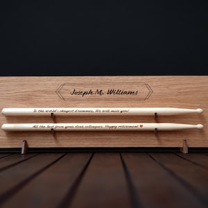 May include: A wooden plaque with a personalized message for a retiring drummer. The plaque has two slots to hold a pair of drumsticks. The message reads "To the world's okayest drummer. We will miss you! All the best from your dear colleagues. Happy retirement".