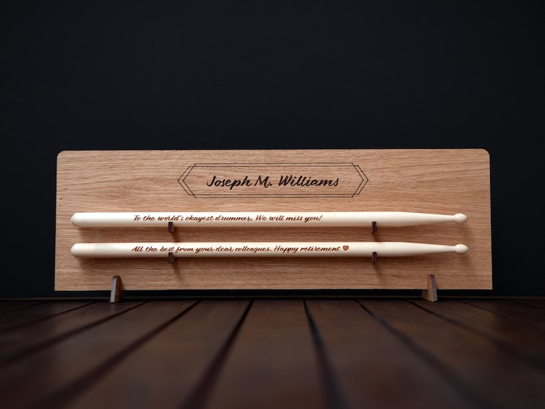 May include: A wooden plaque with a personalized message for a retiring drummer. The plaque has two slots to hold a pair of drumsticks. The message reads "To the world's okayest drummer. We will miss you! All the best from your dear colleagues. Happy retirement".