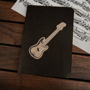 May include: A black notebook with a brown bass guitar sticker on the front. The notebook is lying on a wooden surface.