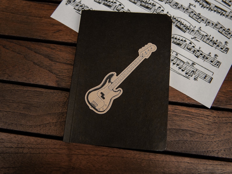 May include: A black notebook with a brown bass guitar sticker on the front. The notebook is lying on a wooden surface.