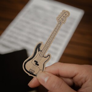 May include: A brown paper cutout of a black bass guitar. The guitar is held in a hand against a white sheet of paper with lines.