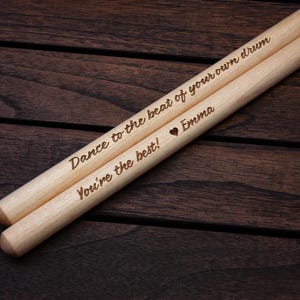 May include: A pair of wooden drumsticks with the text "Dance to the beat of your own drum" and "You're the best!"  The second drumstick also has a heart symbol and the name "Emma" engraved on it.