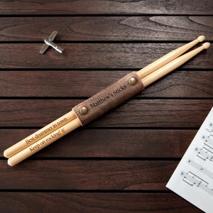 Könnte beinhalten: Ein Paar hölzerne Drumsticks mit den Gravuren "Best drummer in town" und "Keep on rocking!". Die Drumsticks werden in einem braunen Lederetui mit der Aufschrift "Matthew's Sticks" gehalten. Ein Stimmschlüssel und Noten sind ebenfalls zu sehen.