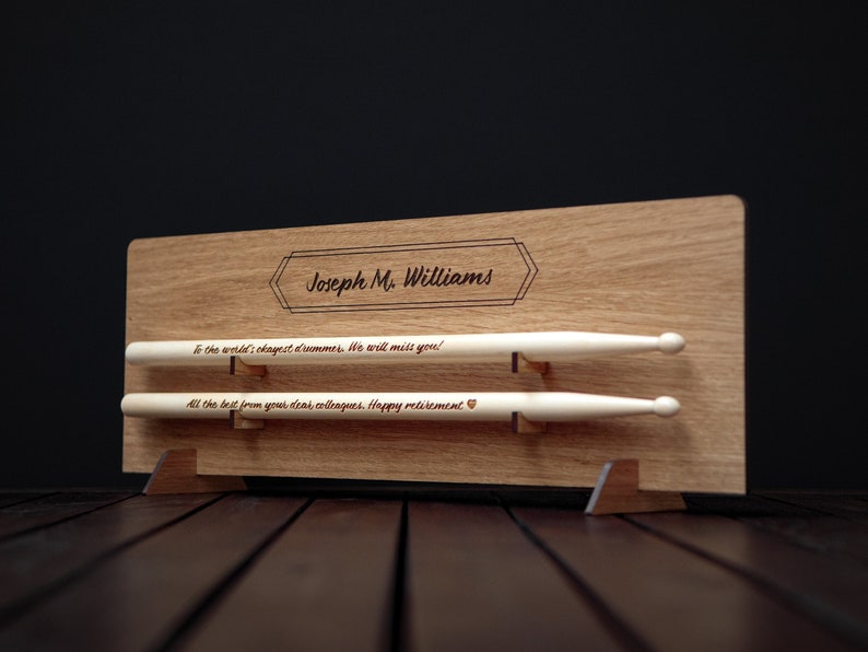 May include: A wooden plaque with a personalized message for a retiring drummer. The plaque has two slots for drumsticks, which are included. The message reads "To the world's okayest drummer. We will miss you! All the best from your dear colleagues. Happy retirement."