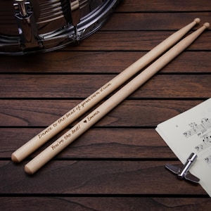 May include: A pair of wooden drumsticks with the text "Dance to the beat of your own drum" and "You're the best! Emma" engraved on them. The drumsticks are lying on a wooden surface next to a sheet of music.