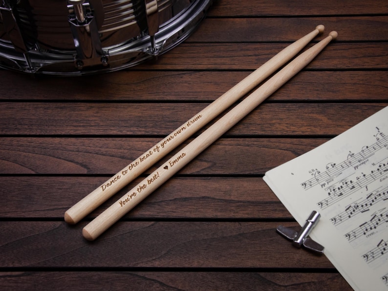May include: A pair of wooden drumsticks with the text "Dance to the beat of your own drum" and "You're the best! Emma" engraved on them. The drumsticks are lying on a wooden surface next to a sheet of music.