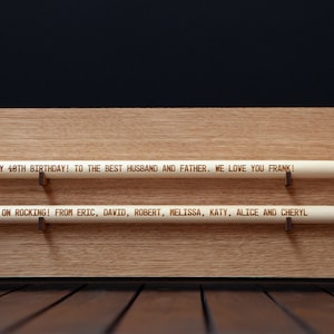 May include: A wooden plaque with two engraved drumsticks. The top drumstick reads "HAPPY 48TH BIRTHDAY! TO THE BEST HUSBAND AND FATHER. WE LOVE YOU FRANK!" The bottom drumstick reads "KEEP ON ROCKING! FROM ERIC, DAVID, ROBERT, MELISSA, KATY, ALICE AND CHERYL"