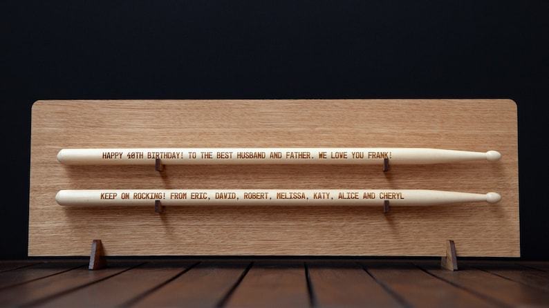 May include: A wooden plaque with two engraved drumsticks. The top drumstick reads "HAPPY 48TH BIRTHDAY! TO THE BEST HUSBAND AND FATHER. WE LOVE YOU FRANK!" The bottom drumstick reads "KEEP ON ROCKING! FROM ERIC, DAVID, ROBERT, MELISSA, KATY, ALICE AND CHERYL"