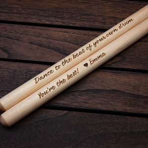 May include: Two wooden drumsticks with the text "Dance to the beat of your own drum" and "You're the best!"  with a heart symbol and the name "Emma" engraved on them.