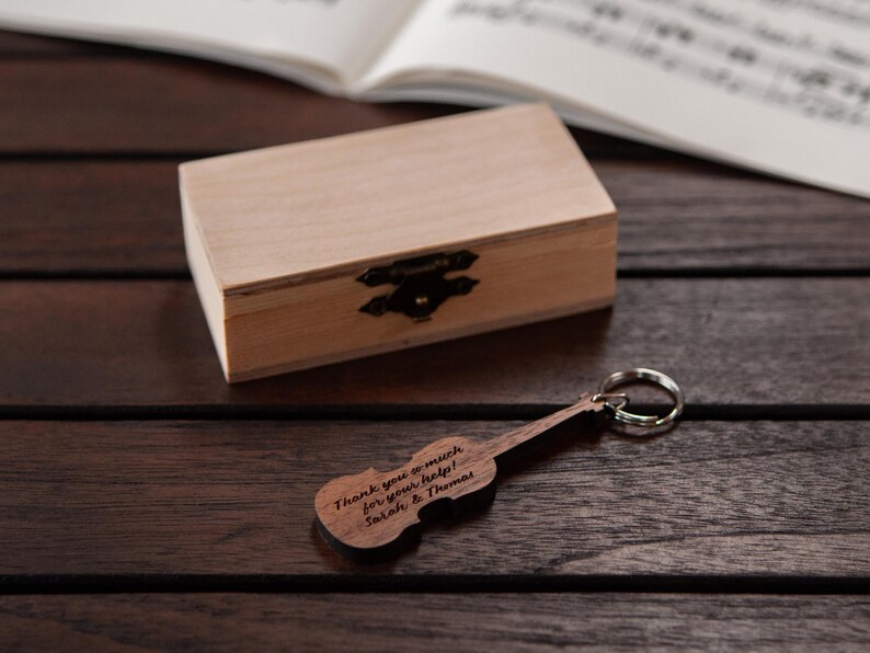 Puede incluir: Un llavero de viol&iacute;n de madera con la inscripci&oacute;n "Thank you so much for your help! Sarah & Thomas" sobre un fondo de madera con una peque&ntilde;a caja de madera.