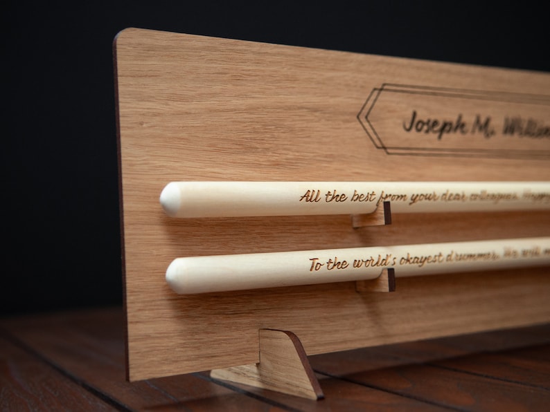 May include: A wooden plaque with a personalized message for Joseph M. Williams. The plaque has two wooden dowels with engraved messages: "All the best from your dear colleagues" and "To the world's okayest drummer".