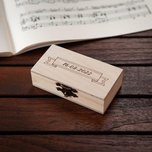 Puede incluir: Una peque&ntilde;a caja de madera con tapa con bisagras y cierre de lat&oacute;n. La caja est&aacute; grabada con la fecha "16-03-2022" y una pancarta decorativa.