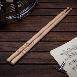 May include: A pair of wooden drumsticks with the text "No Drums, No Life" and "Know Drums, Know Life" engraved on them. The drumsticks are lying on a wooden surface next to a sheet of music and a drum.