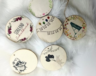 Peut inclure: Six décorations rondes en bois avec des motifs différents. Une décoration a un motif floral avec le texte "Schulkind Schulkind". Une autre décoration a un motif floral avec la lettre "A" et le texte "Amelie". Une autre décoration a un motif de dinosaure. Une autre décoration a un motif avec des ballons et le texte "2024". Une autre décoration a un motif avec des ballons et le texte "Schulkind". La dernière décoration est vide.