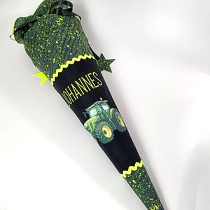 Puede incluir: Un cono escolar verde y negro con un gráfico de tractor. El cono presenta el nombre "JOHANNES" en letras amarillas. La parte superior del cono es verde con motas amarillas y una cinta negra. El borde inferior está adornado con un patrón en zigzag amarillo.