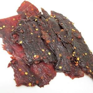 May include: Close-up of a pile of dark red beef jerky with a spicy, peppery seasoning. The jerky is arranged in a random pattern, with some pieces overlapping.