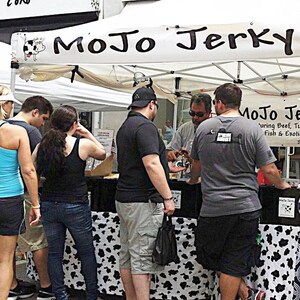 May include: A street food vendor selling jerky under a white canopy tent with the sign "MoJo Jerky" featuring beef, turkey, fish, and exotic meats.