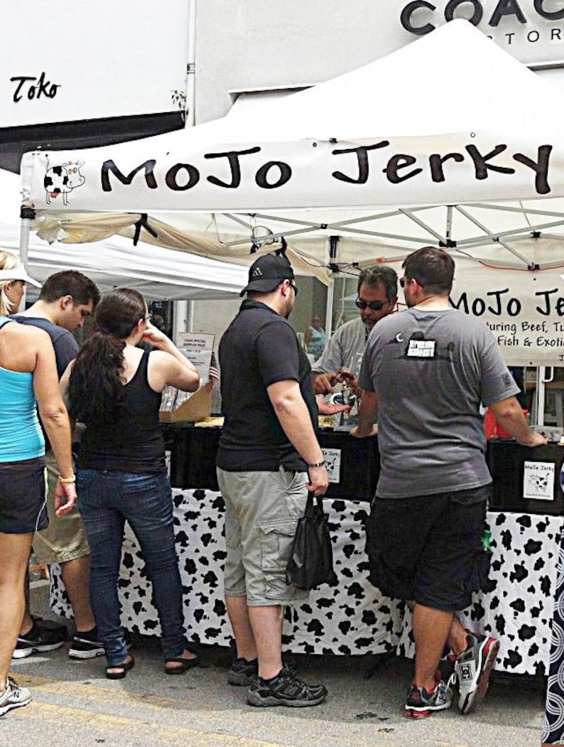 May include: A street food vendor selling jerky under a white canopy tent with the sign "MoJo Jerky" featuring beef, turkey, fish, and exotic meats.